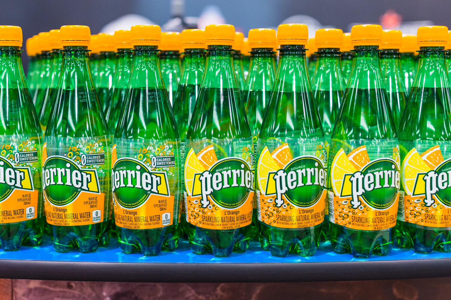 Dense rows of green Perrier L'Orange bottles with yellow caps on a blue-lit shelf.