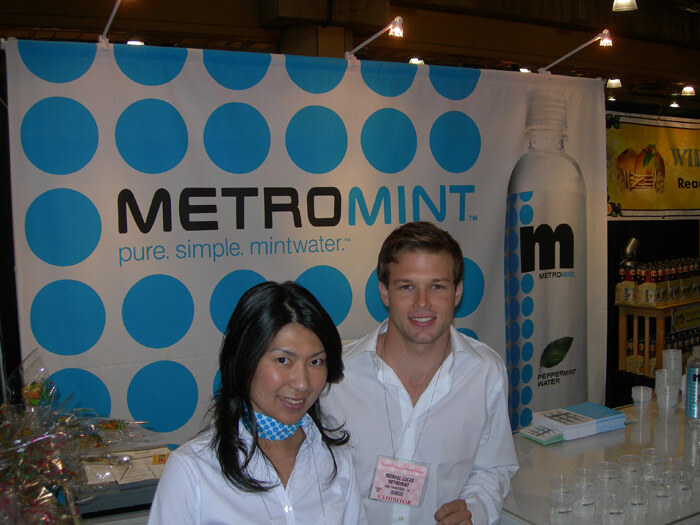 A dark-haired woman in a white top and blue scarf and a man in a white shirt with an exhibitor badge pose in front of a blue-dotted MetroMint banner reading pure. simple. mintwater.