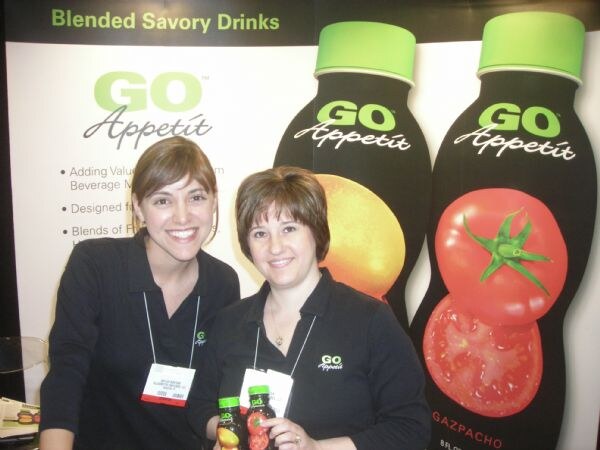 Two smiling women in black Go Appetit shirts and lanyards pose with small bottles in front of Go Appetit banners reading Blended Savory Drinks, including a gazpacho bottle graphic.