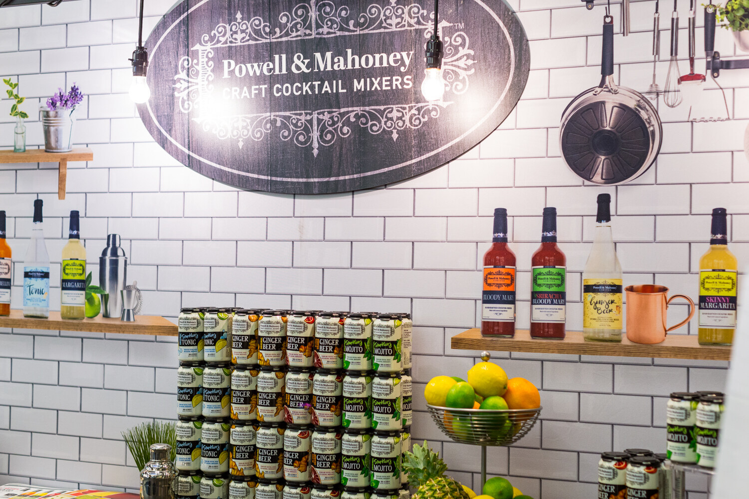 Booth wall with an oval Powell & Mahoney Craft Cocktail Mixers sign, shelves of mixer bottles, a copper mug and stacked ginger beer and mojito cans.