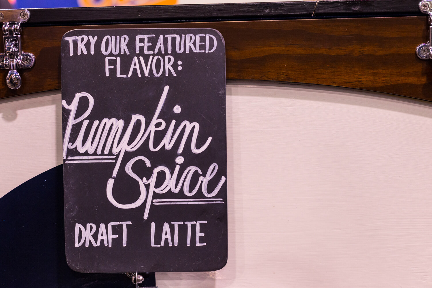 Black chalkboard sign reading Try our featured flavor: Pumpkin Spice Draft Latte on a cream wall.
