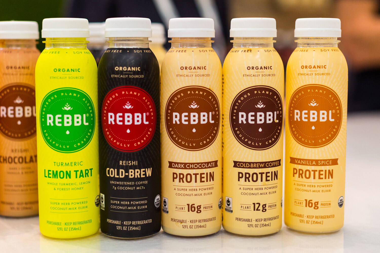Row of Rebbl organic bottles including Turmeric Lemon Tart, Reishi Cold-Brew and three protein flavors.