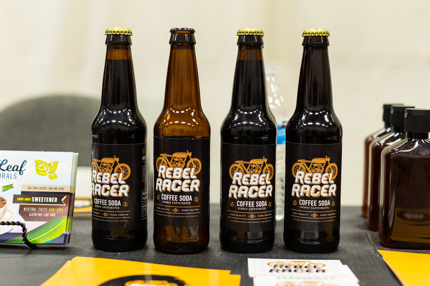 Four glass bottles labeled Rebel Racer Coffee Soda with a motorcycle logo, with brochures on the table.