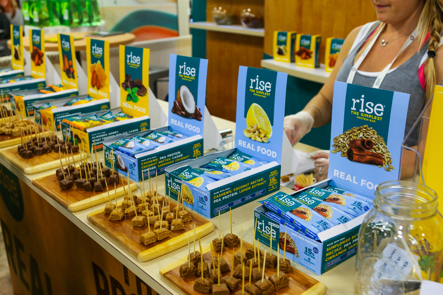 Counter with boxes of Rise protein bars, display cards and boards of skewered bar samples, with a woman behind.