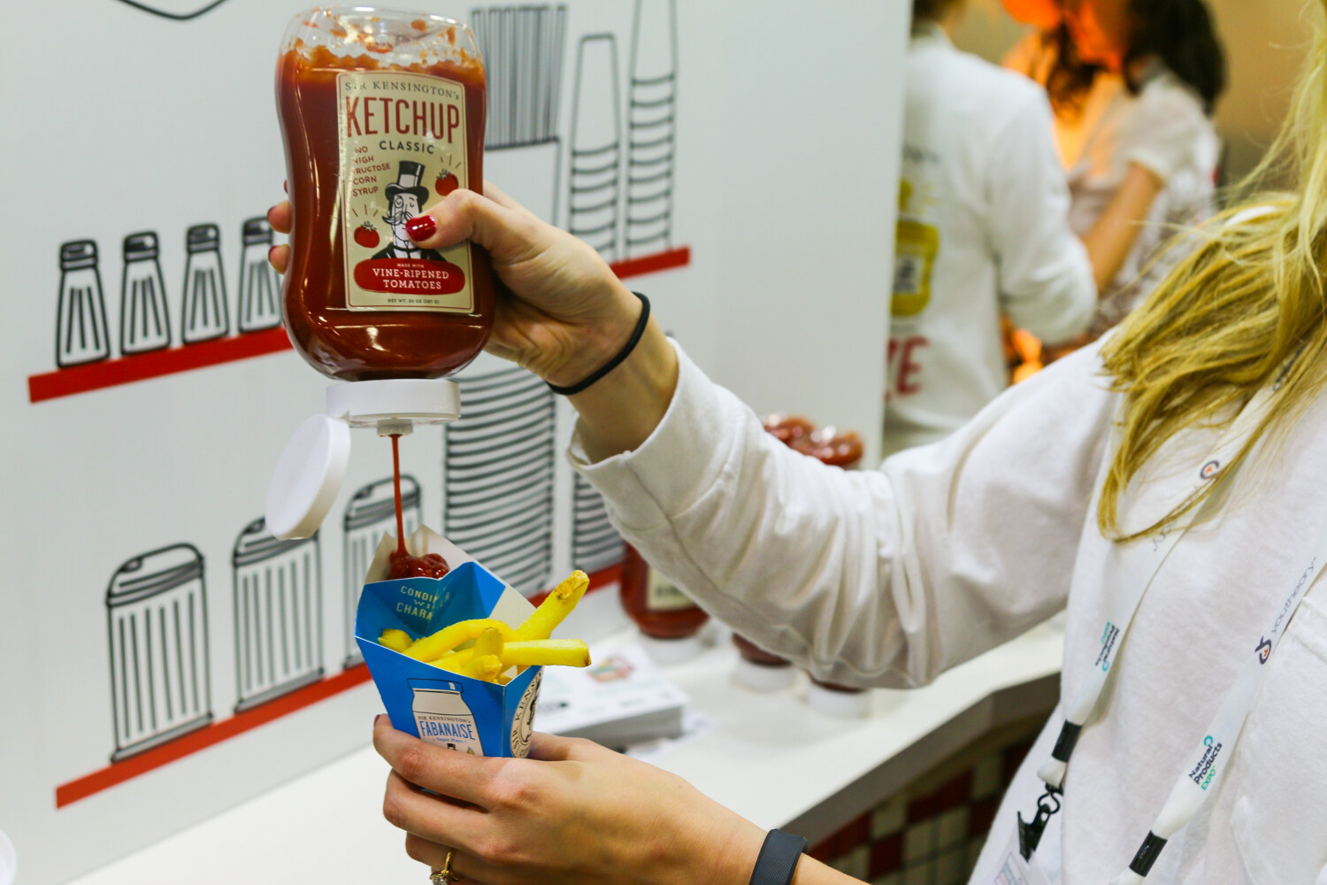 Hand squeezing a bottle of Sir Kensington's Classic ketchup onto french fries in a blue paper cone.