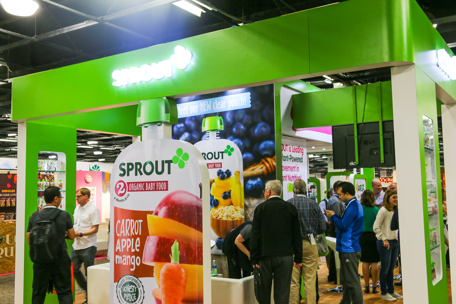 Large green Sprout booth with a giant Carrot Apple Mango pouch graphic and a crowd of attendees on the right.