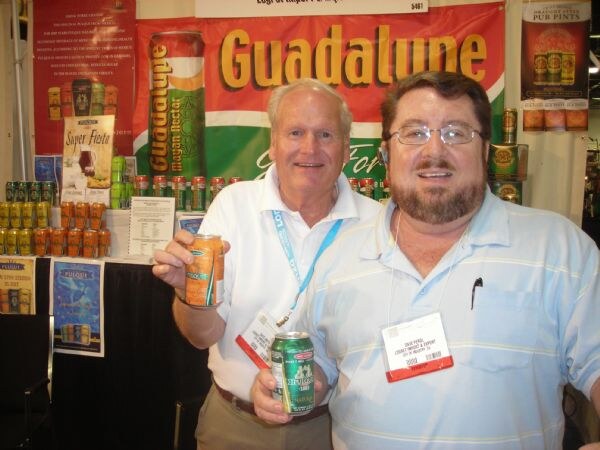 Two men stand in front of a Guadalupe banner, one in a white polo holding up a can and the other in a blue striped shirt and glasses holding a green can. Cans are stacked behind them.