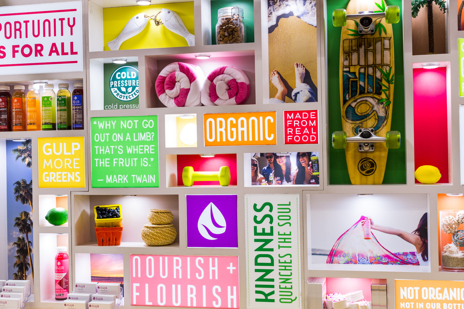 Grid of lit shelves with signs such as Organic and Gulp More Greens, a skateboard, towels, fruit and Suja bottles.
