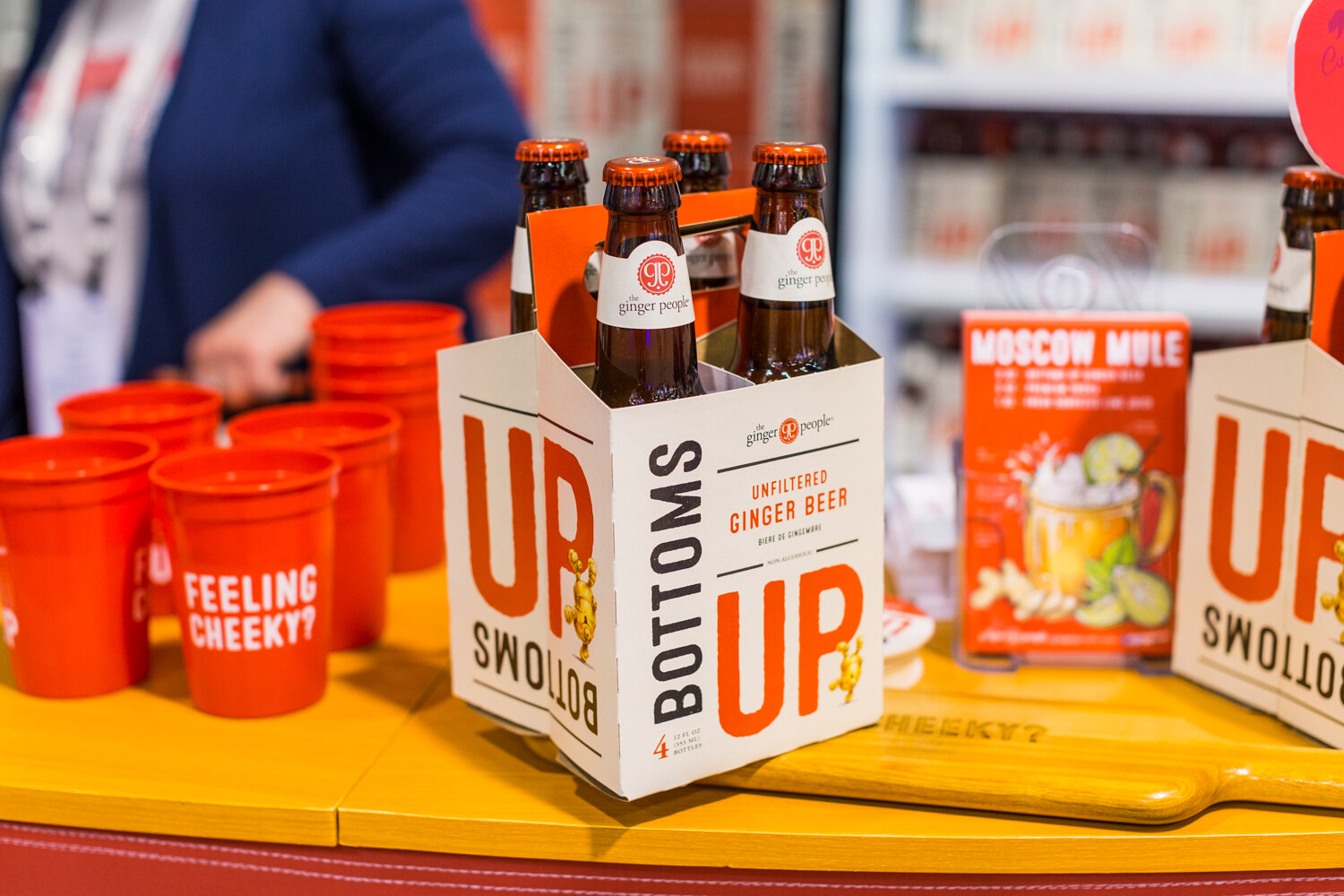 Four-bottle carrier labeled Bottoms Up Unfiltered Ginger Beer by The Ginger People, with orange cups reading Feeling Cheeky and a Moscow Mule card.