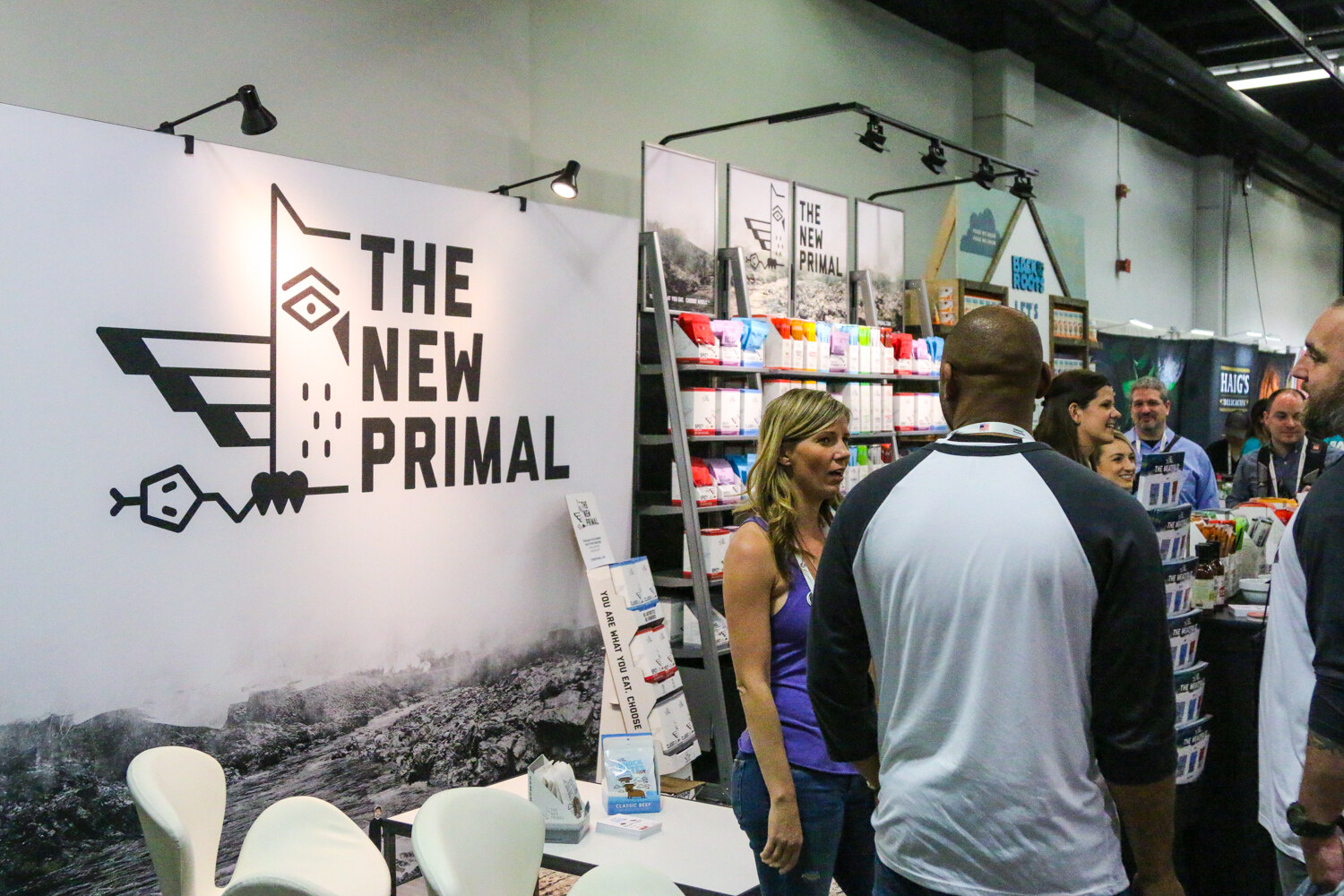Booth with a large The New Primal owl logo wall, shelves of jerky packs and several people talking in the aisle.