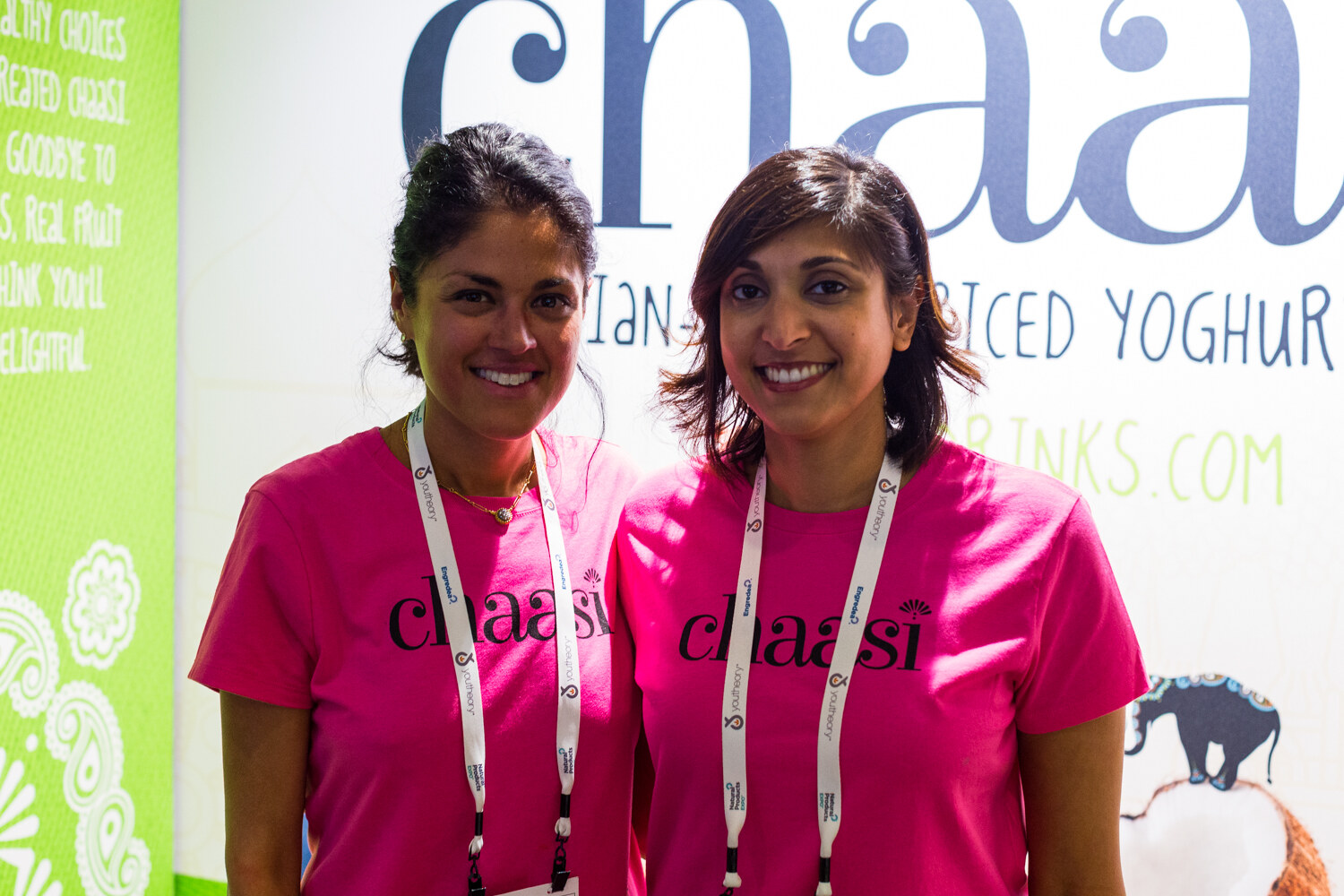 Two smiling women in pink Chaasi T-shirts and lanyards standing in front of a Chaasi banner.
