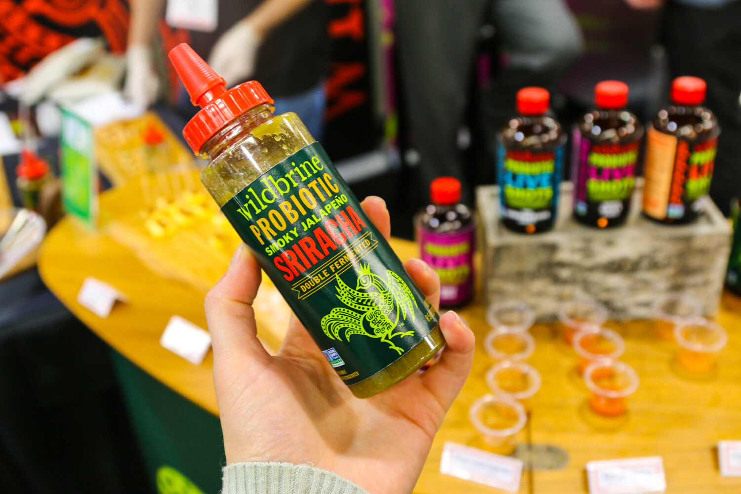 Hand holding a Wildbrine Probiotic Smoky Jalapeno Sriracha bottle with other Wildbrine bottles and sample cups behind.