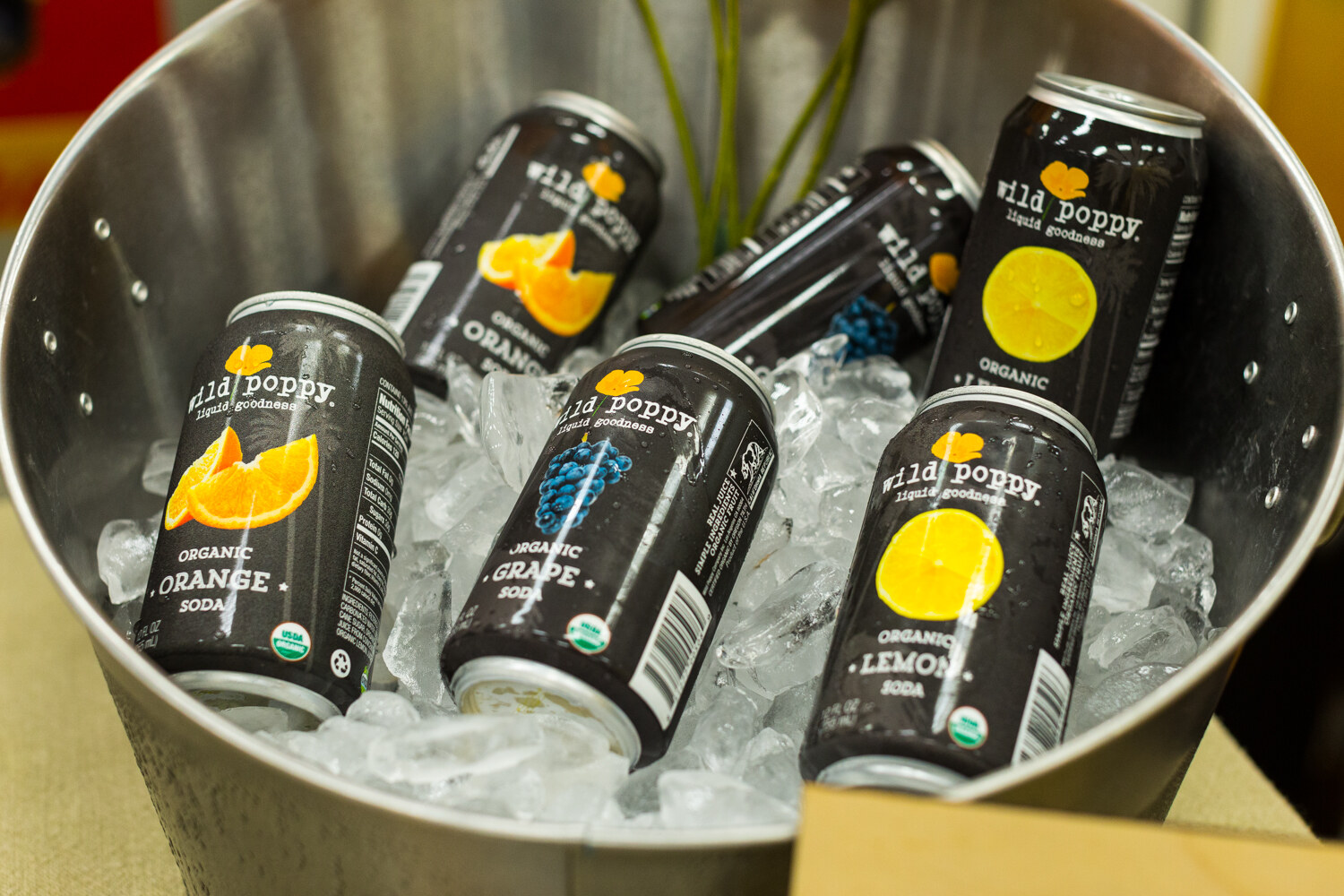 Silver tub filled with ice and cans of Wild Poppy organic orange, grape and lemon soda.
