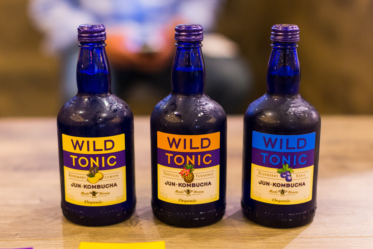 Three cobalt blue bottles labeled Wild Tonic Jun Kombucha in rosemary lemon, tropical turmeric and blueberry basil.