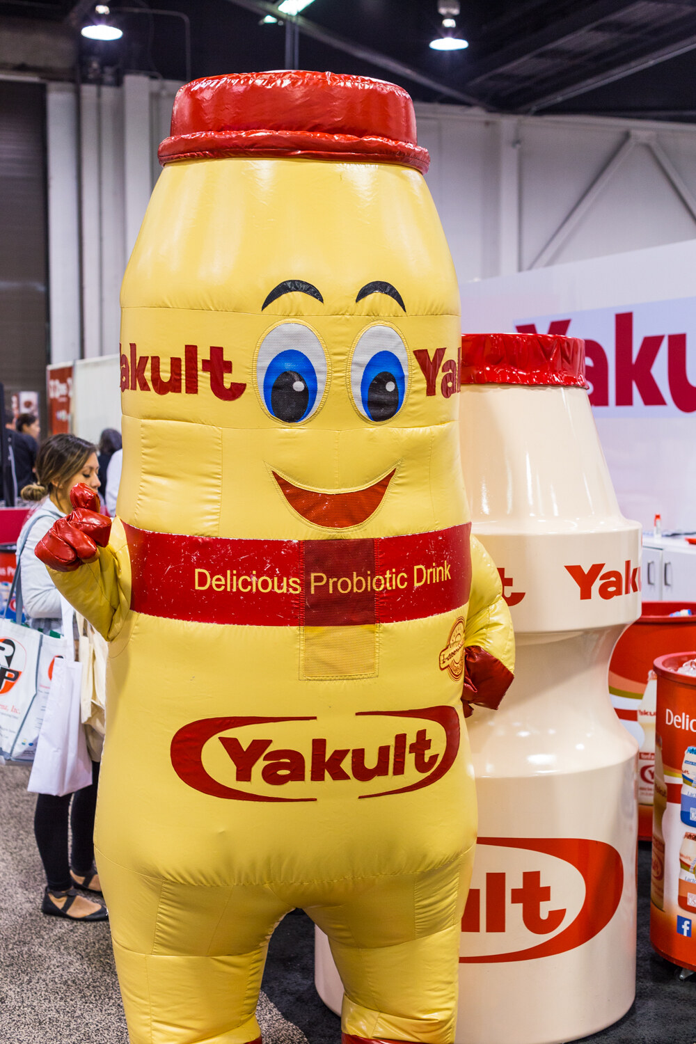Person in an inflatable yellow Yakult bottle costume with a smiling face, beside a large Yakult bottle prop.