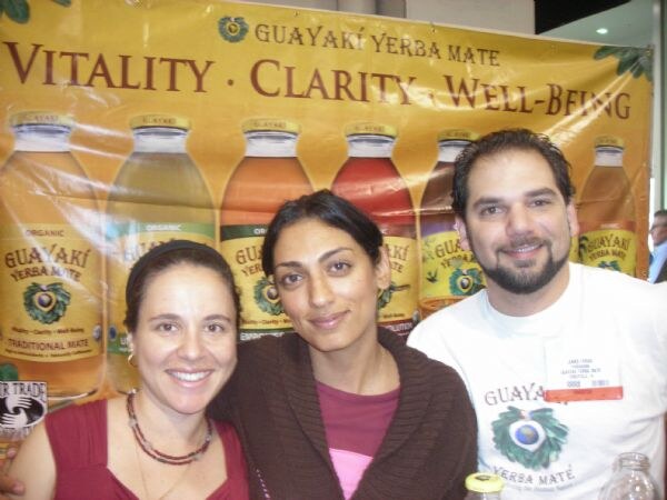 Two women and a bearded man smile in front of a Guayakí Yerba Mate banner reading Vitality, Clarity, Well-Being, with bottles of the drink pictured behind them.