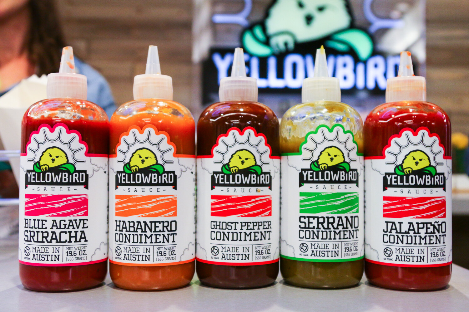 Five squeeze bottles of Yellowbird sauces including Blue Agave Sriracha and Habanero Condiment in front of a neon Yellowbird sign.