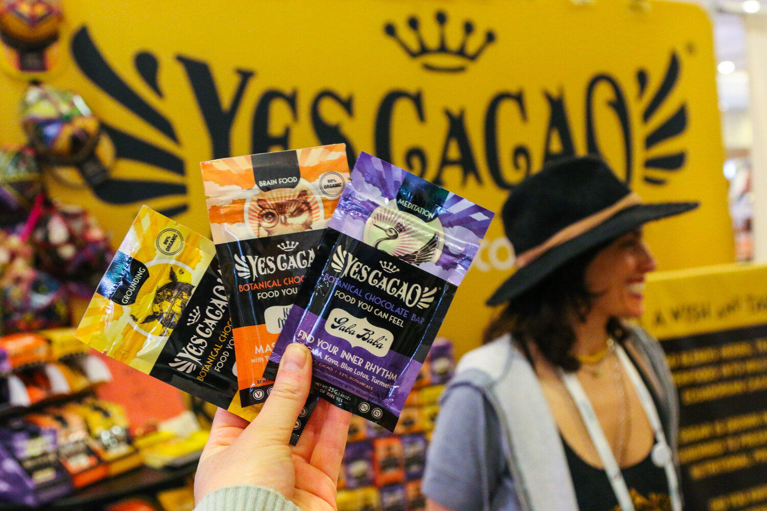 Hand holding three Yes Cacao botanical chocolate bars in front of a yellow Yes Cacao sign and a woman in a black hat.