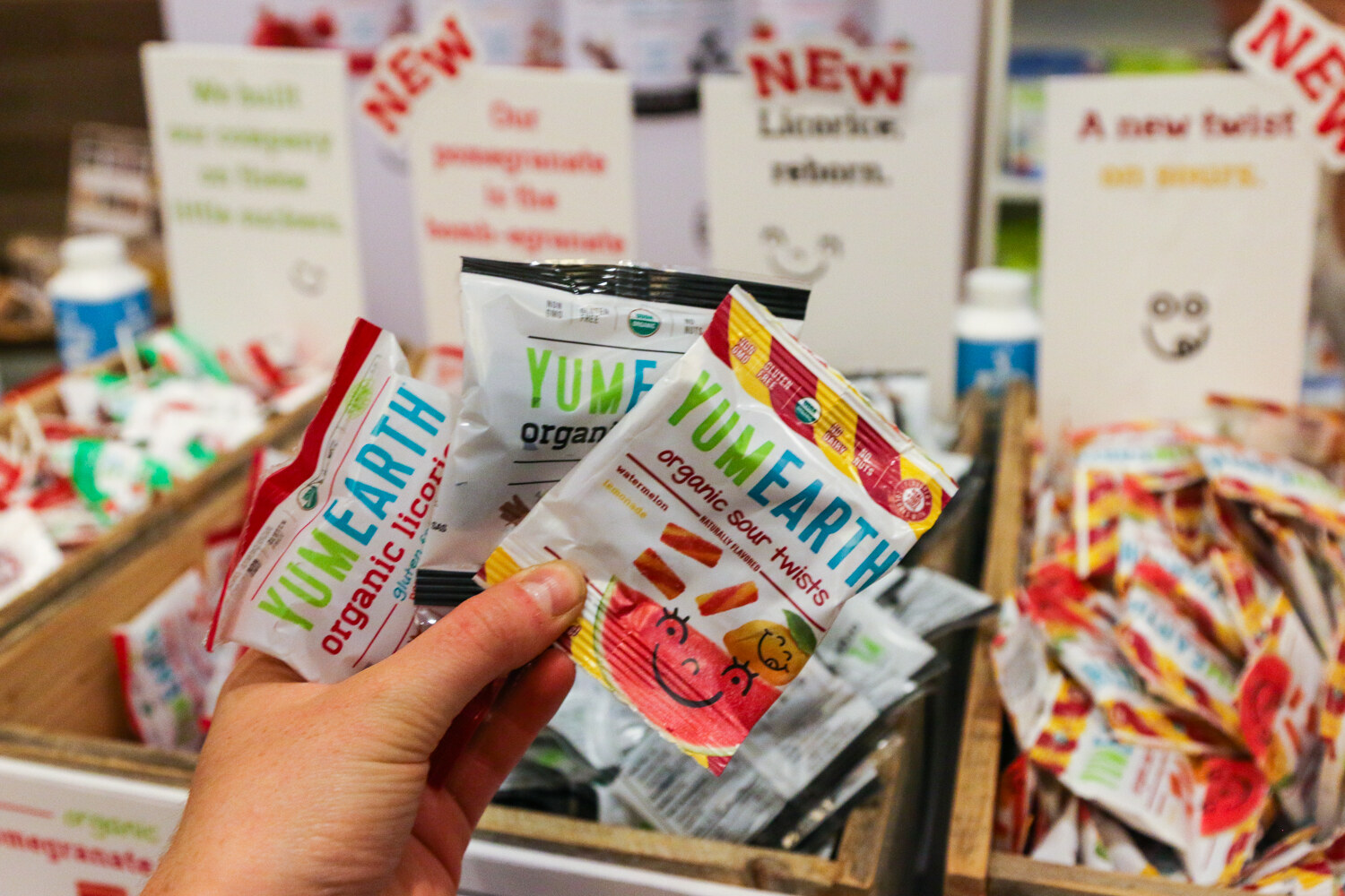 Hand holding several small YumEarth organic candy packets, with open crates of more packets and product signs behind.