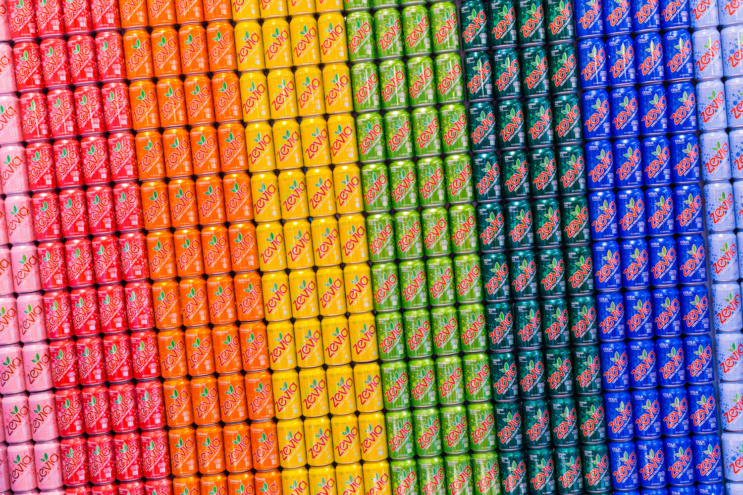 Dense wall of stacked Zevia cans arranged in color bands from pink to orange, yellow, green, blue and light blue.