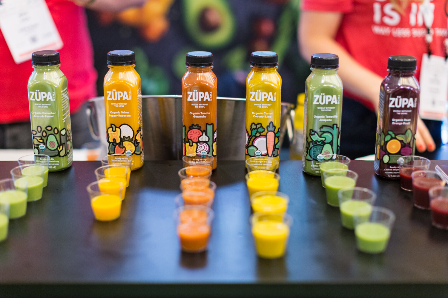 Six bottles of Zupa Noma organic soups lined up on a black table with small cups of green, yellow, orange and red soup in front.