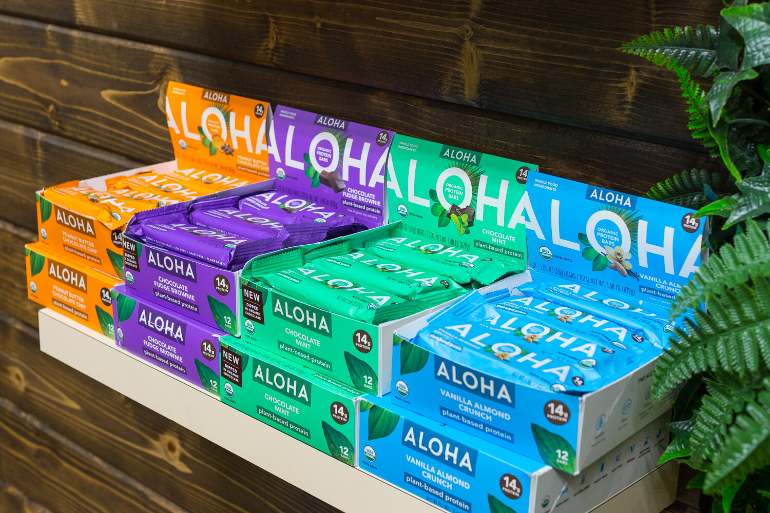 Wall shelf stacked with colorful Aloha protein bar boxes against a wooden wall and fern.