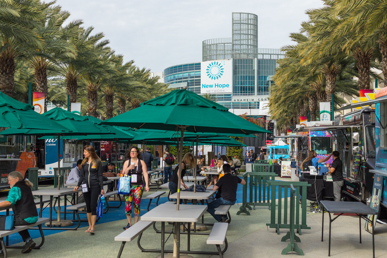 Wide view of outdoor picnic tables with green umbrellas and food trucks lining a palm-tree-shaded plaza, with a glass convention center tower in the background.
