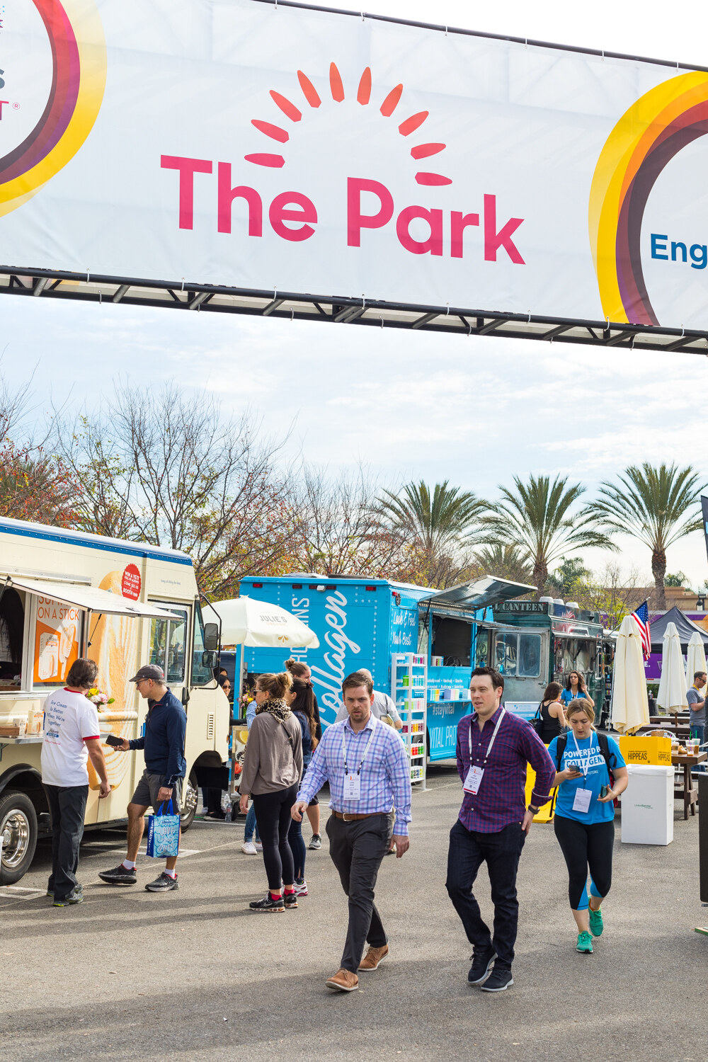 Vertical view of people with badges walking under a banner reading 'The Park' toward a row of food trucks.