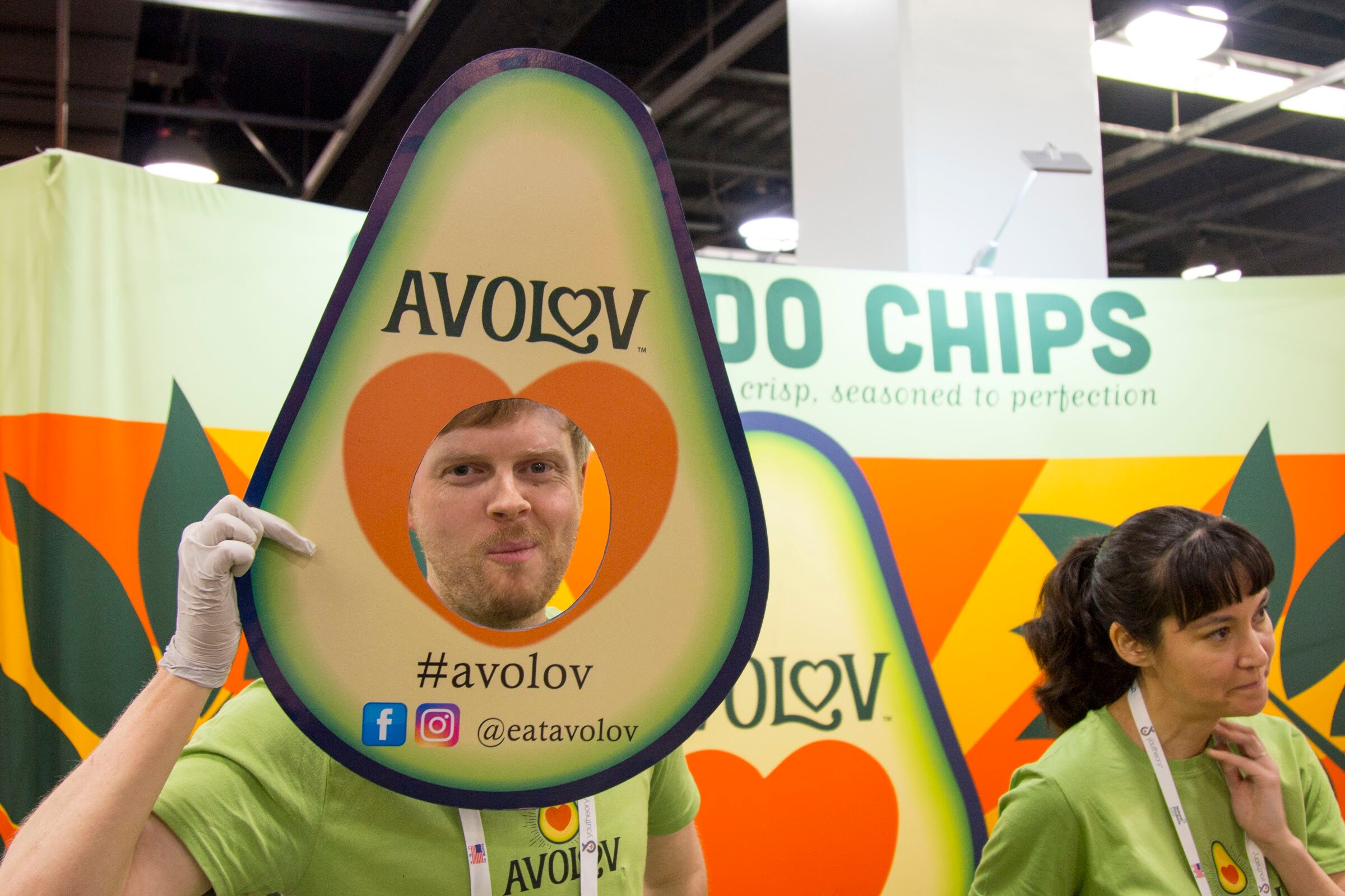 Man in a green Avolov shirt holding an avocado-shaped photo cutout with the hashtag #avolov in front of an avocado chips banner, with a woman beside him.