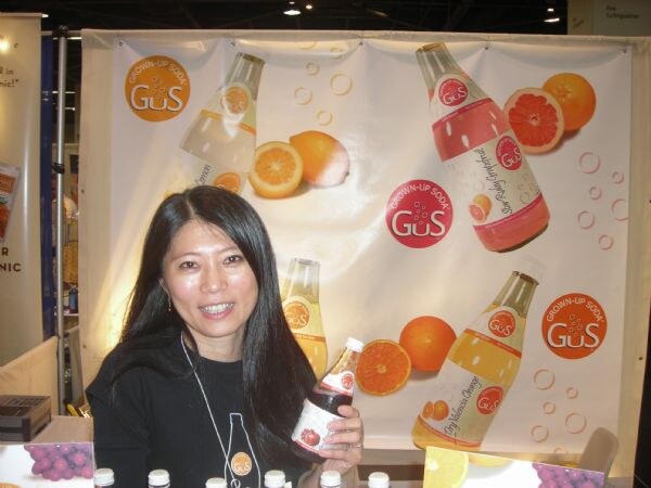 A smiling woman with long dark hair holds a bottle of GuS Grown-Up Soda in front of a banner showing bottles and citrus slices, with more bottles on the table in front.