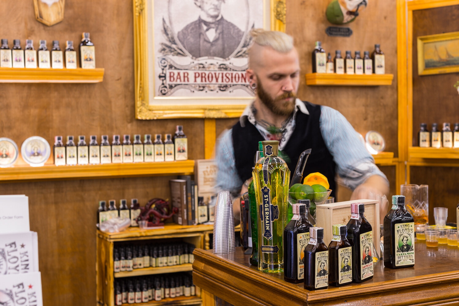 Bearded bartender in a vest working behind a wooden bar with shelves of small bitters bottles and a framed portrait labelled Bar Provisions.