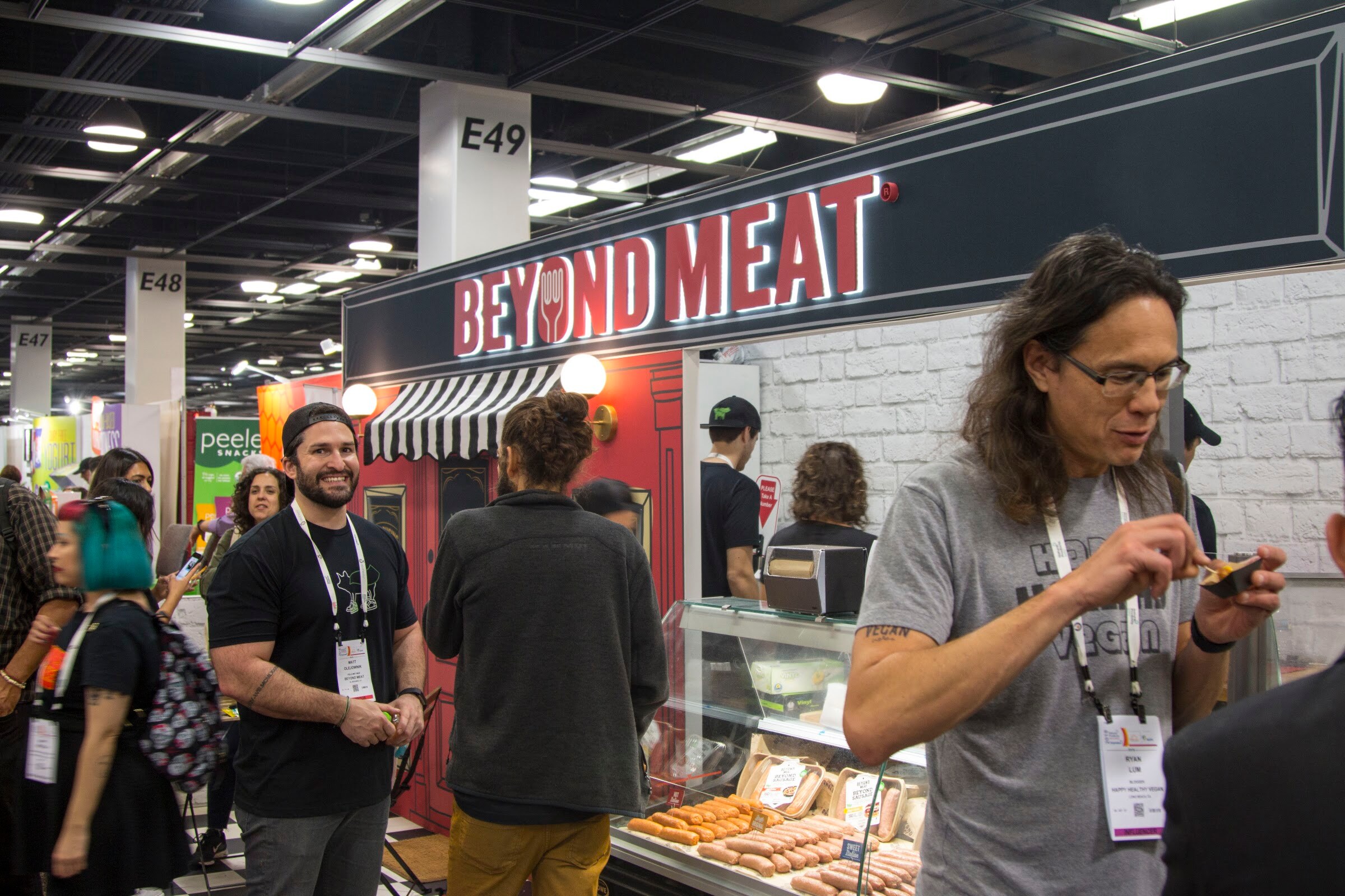 Storefront-style Beyond Meat booth with a lit red sign, a striped awning, a deli case of sausages and attendees chatting in the foreground.