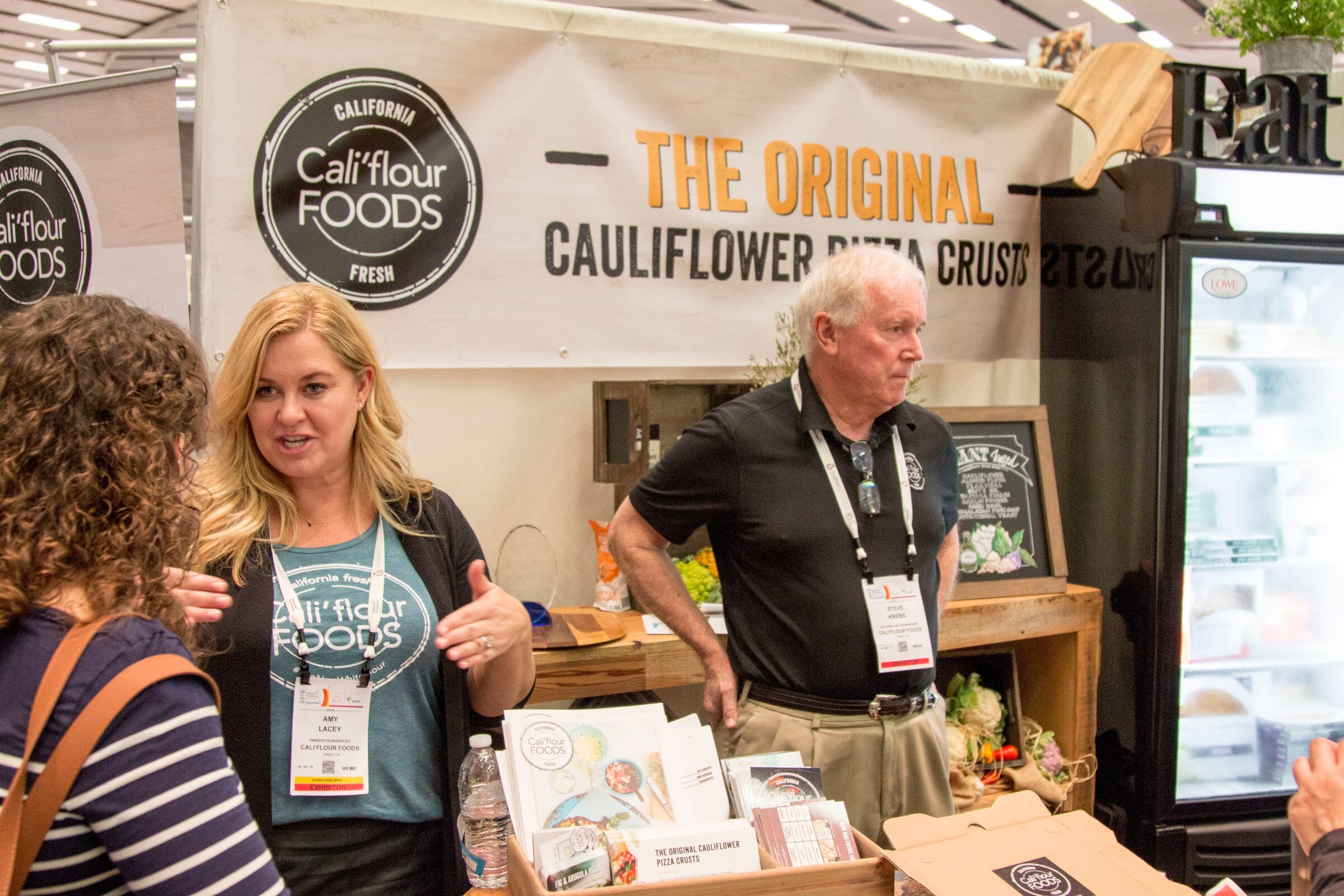 Blond woman in a Cali'flour Foods shirt speaking to a visitor while an older man in a black polo stands beside her, under a banner for cauliflower pizza crusts.