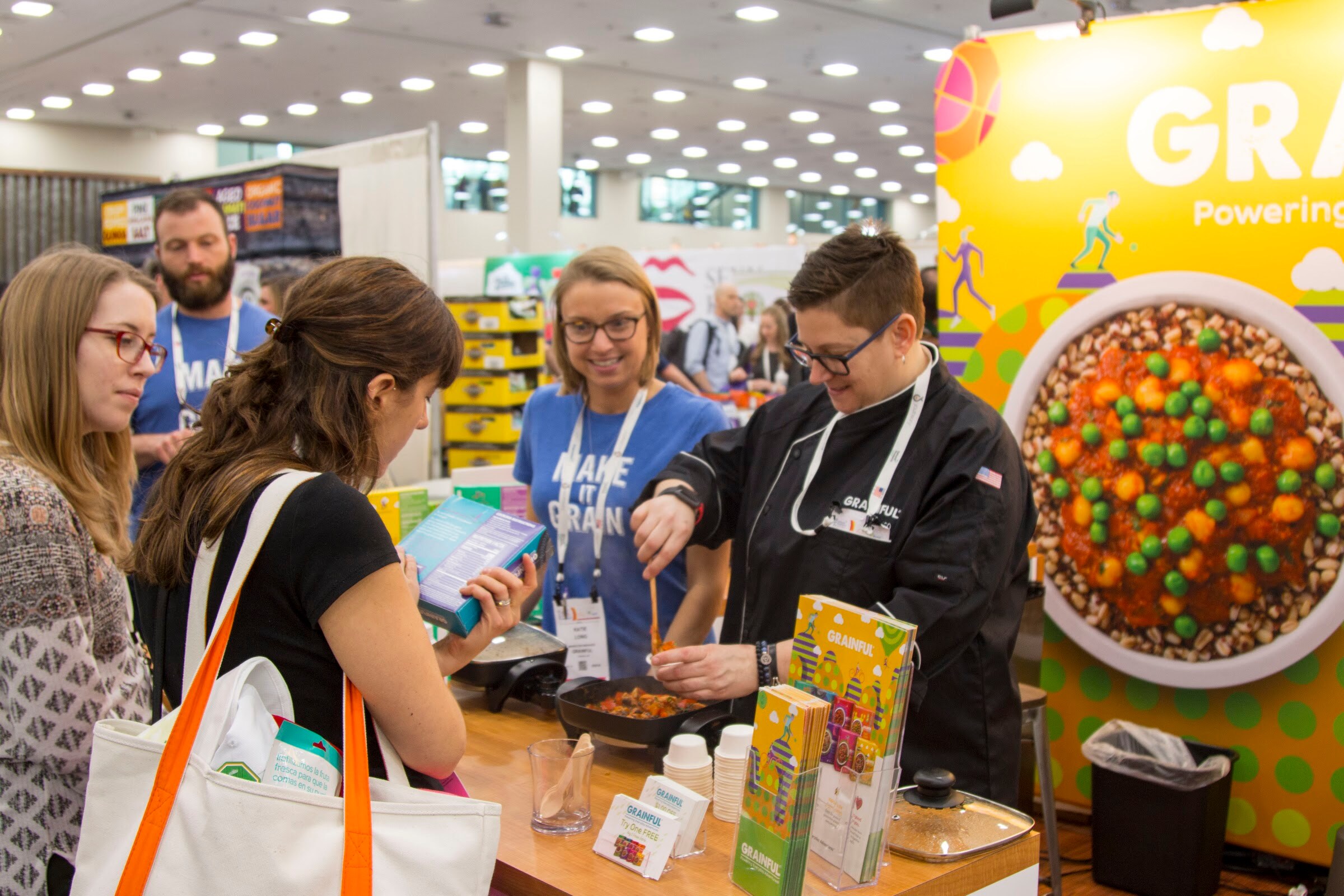 Chef in a black jacket stirring a skillet at a counter while three people watch, in front of a colorful Grainful backdrop.