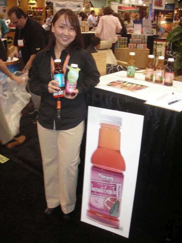 A smiling woman in a black shirt holds two Hansen's tea bottles beside a large Pomegranate Green Tea bottle graphic, with more bottles on a draped table and other booths behind.