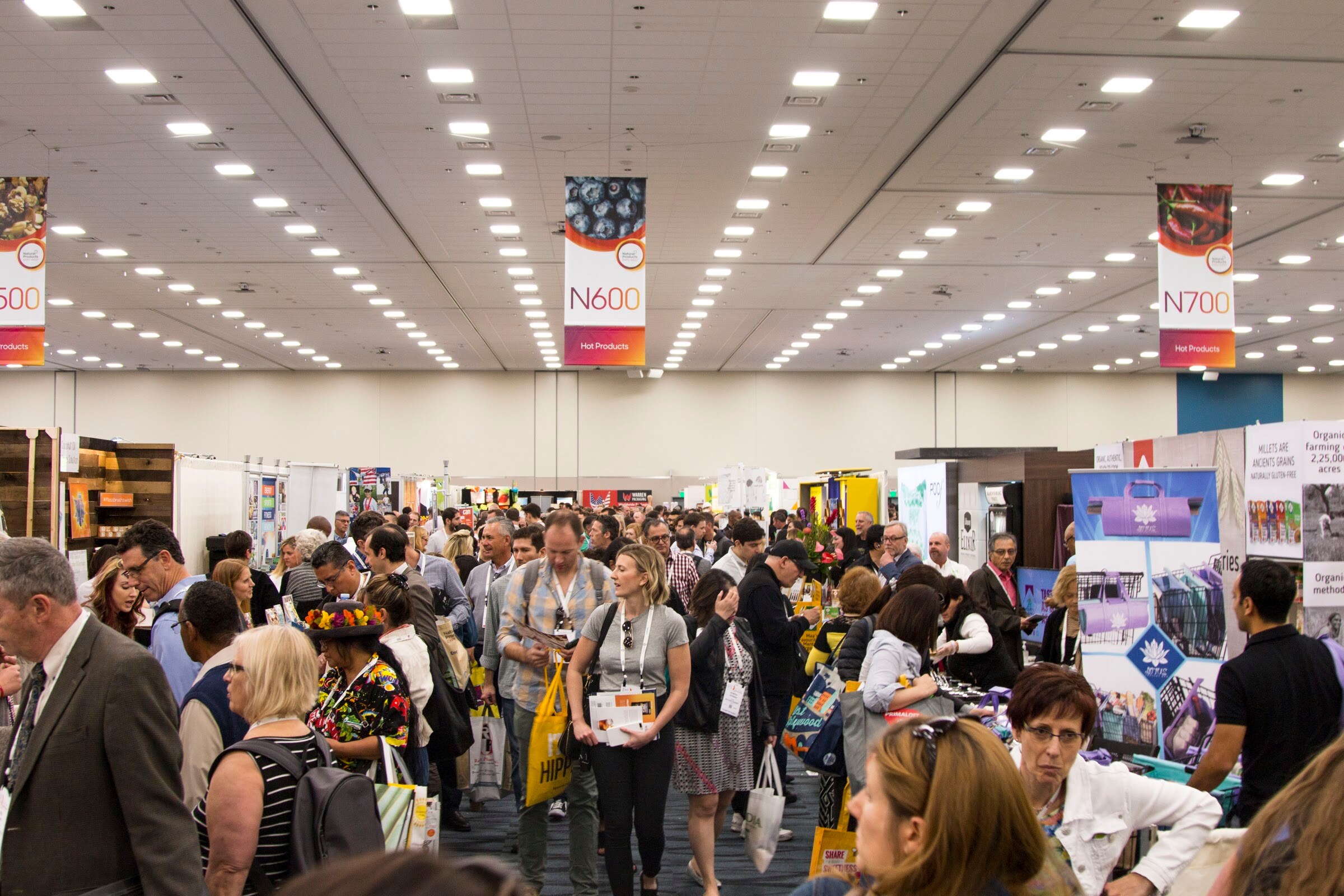 Wide shot of a crowded exhibit hall aisle with attendees carrying bags and overhead hanging banners marked N600 and N700.