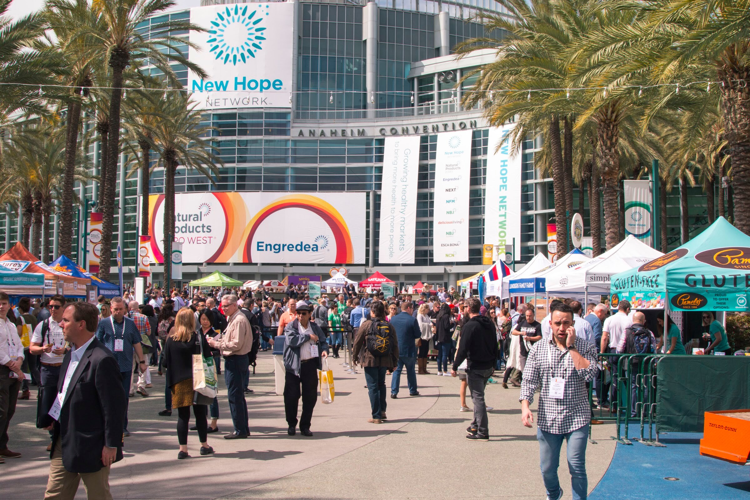Busy outdoor plaza with many attendees walking between white tents, palm trees and a convention center with a large New Hope Network banner.