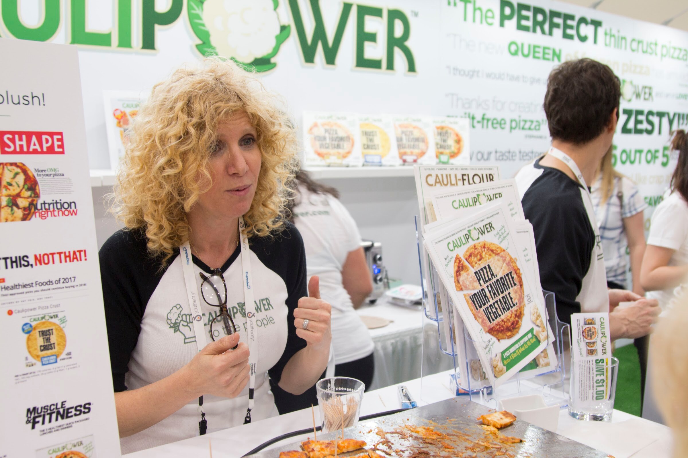 Woman with curly blond hair and a baseball tee talking at a booth with Caulipower signage, product boxes and press clippings behind her.