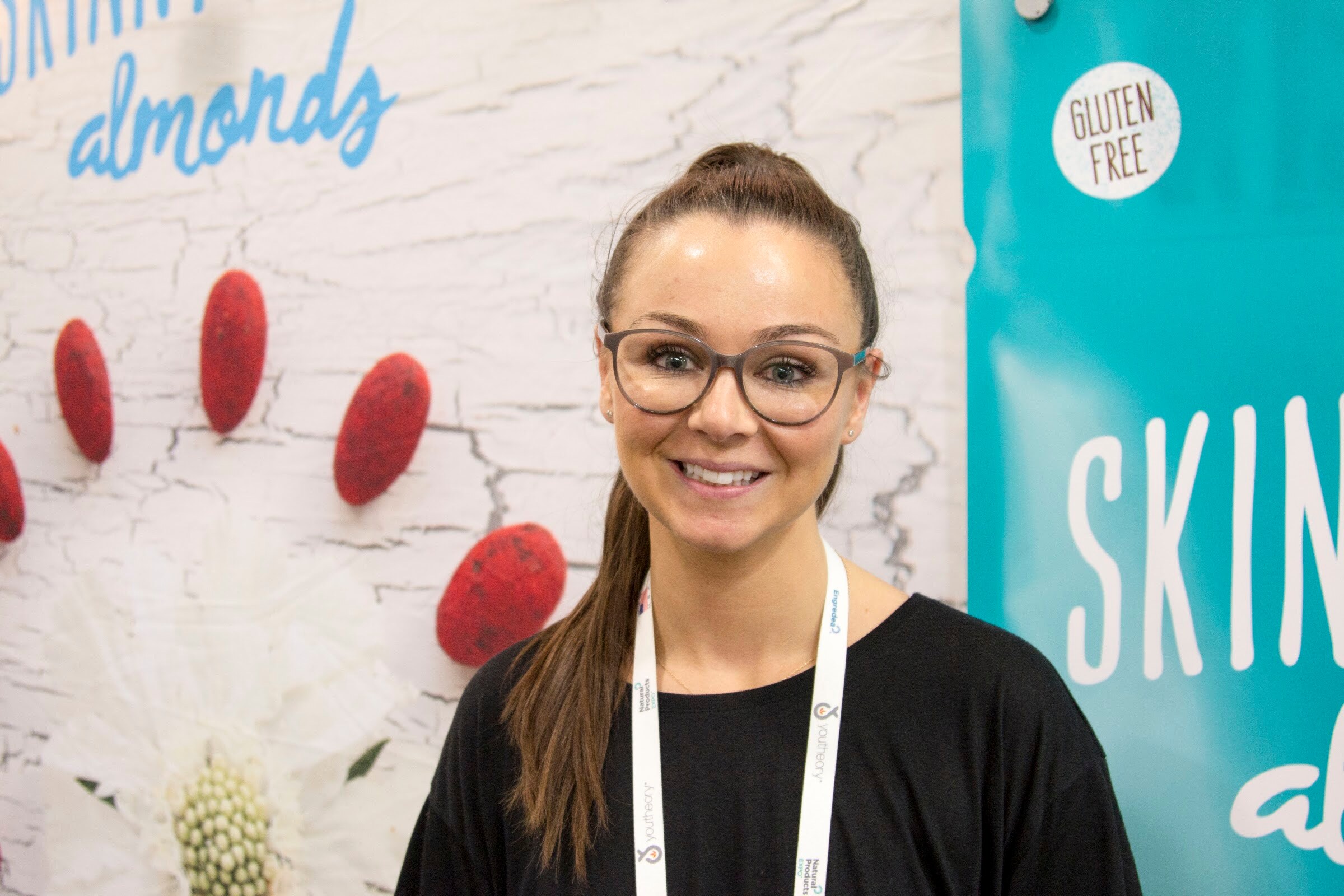 Smiling woman with glasses and a ponytail wearing a lanyard in front of a white wall with printed almonds and the words 'almonds'.