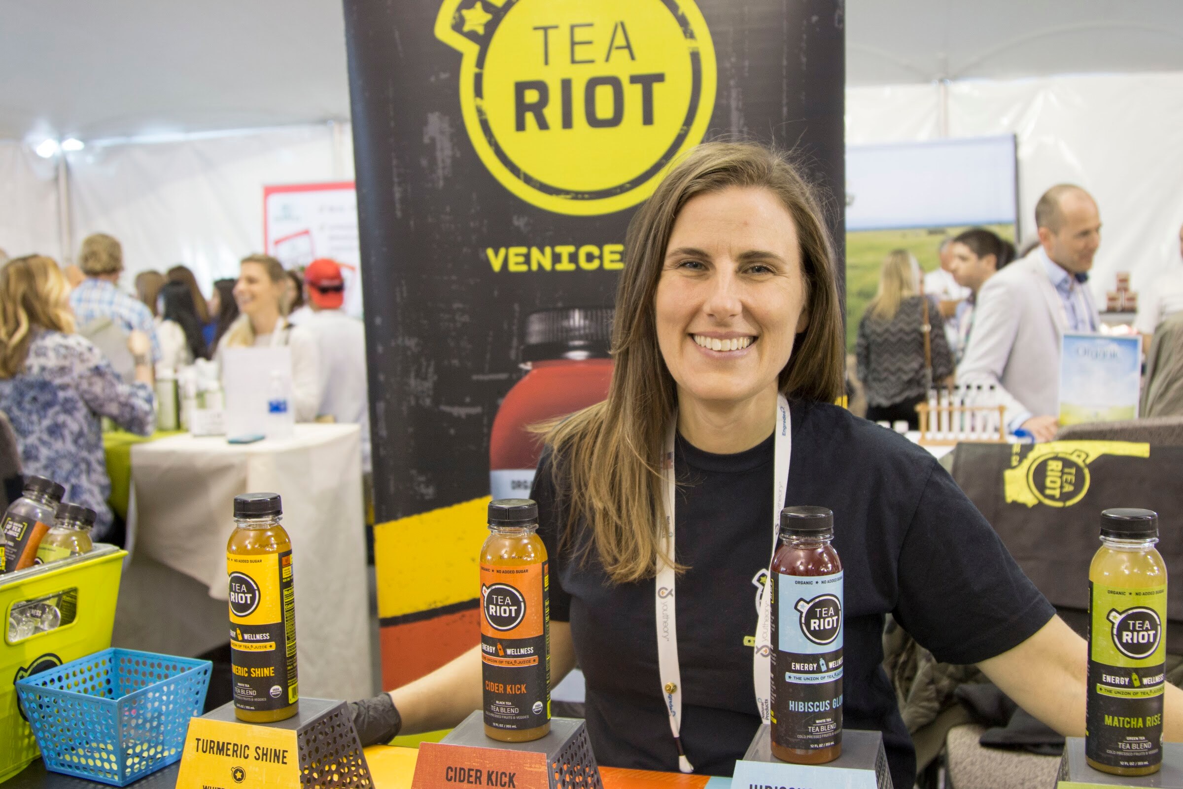 Smiling woman in a black Tea Riot shirt leaning on a counter with bottles of turmeric shine, cider kick and hibiscus tea in front of a Tea Riot banner.