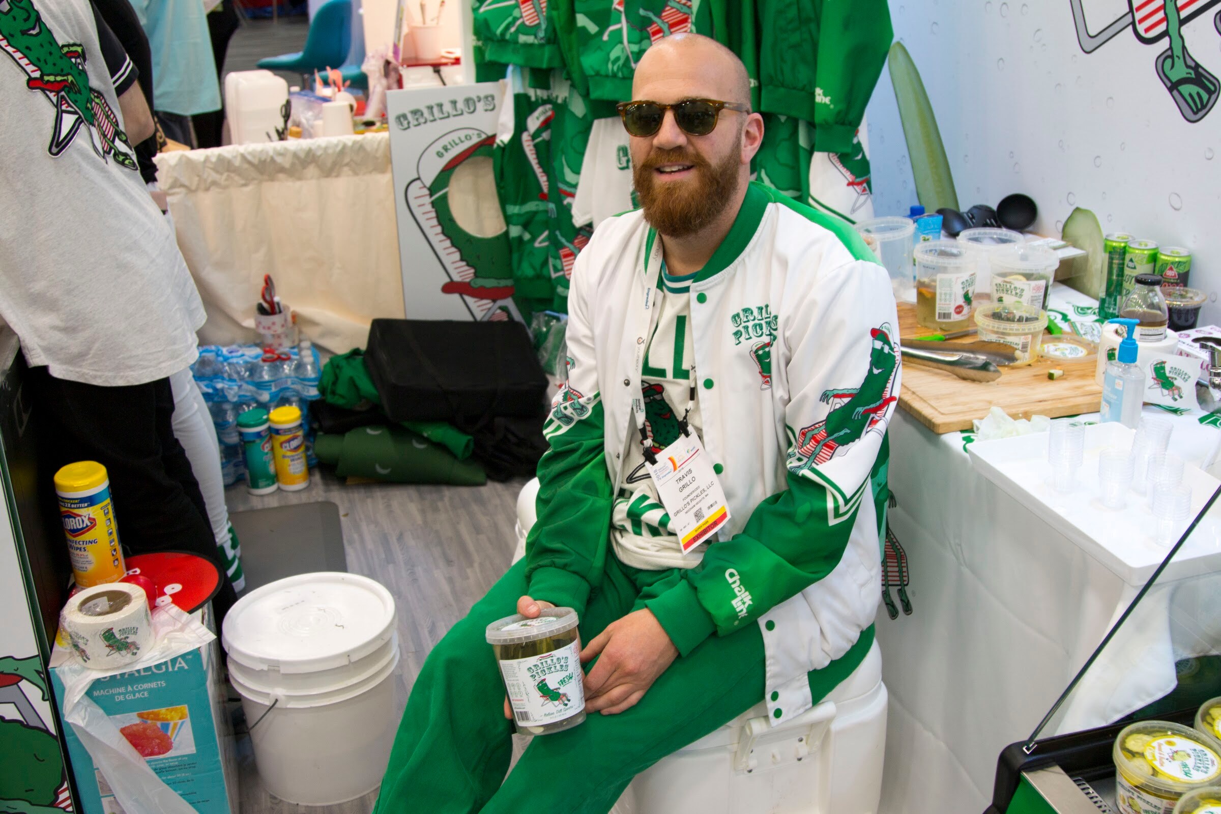 Bearded man in sunglasses and a green and white Grillo's jacket seated in a cluttered booth holding a pickle container.