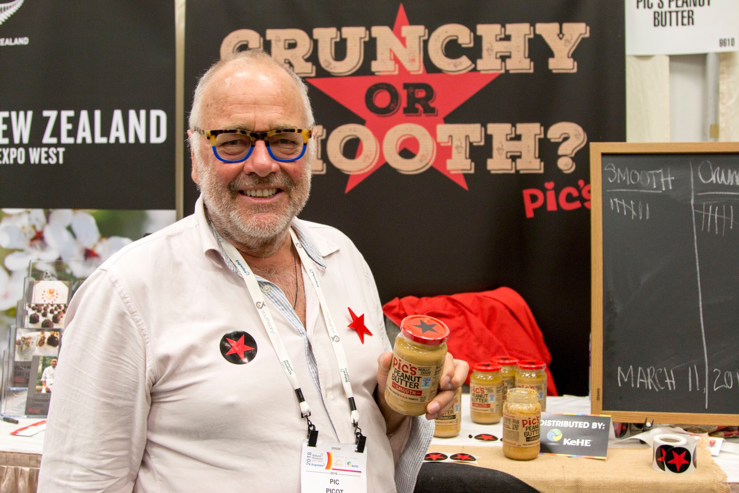 Grey-bearded man in blue-framed glasses and a white shirt holding a jar of Pic's Peanut Butter in front of a sign reading 'Crunchy or Smooth?'.