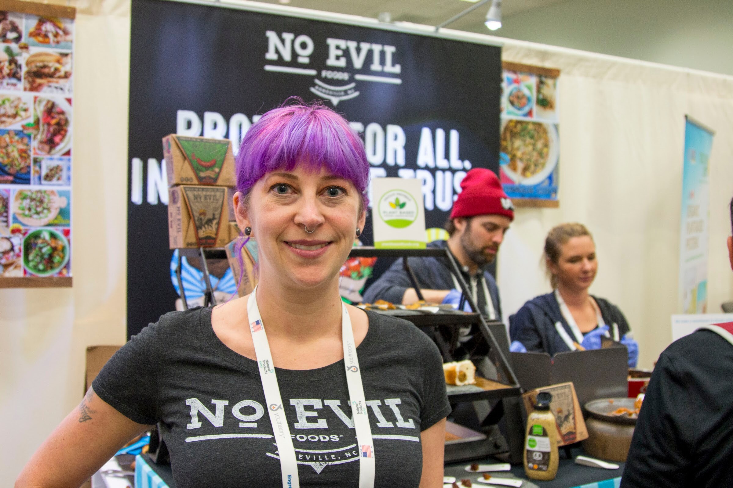 Smiling woman with a purple fringe wearing a No Evil Foods shirt and lanyard in front of a black banner, with two cooks working at a grill behind.