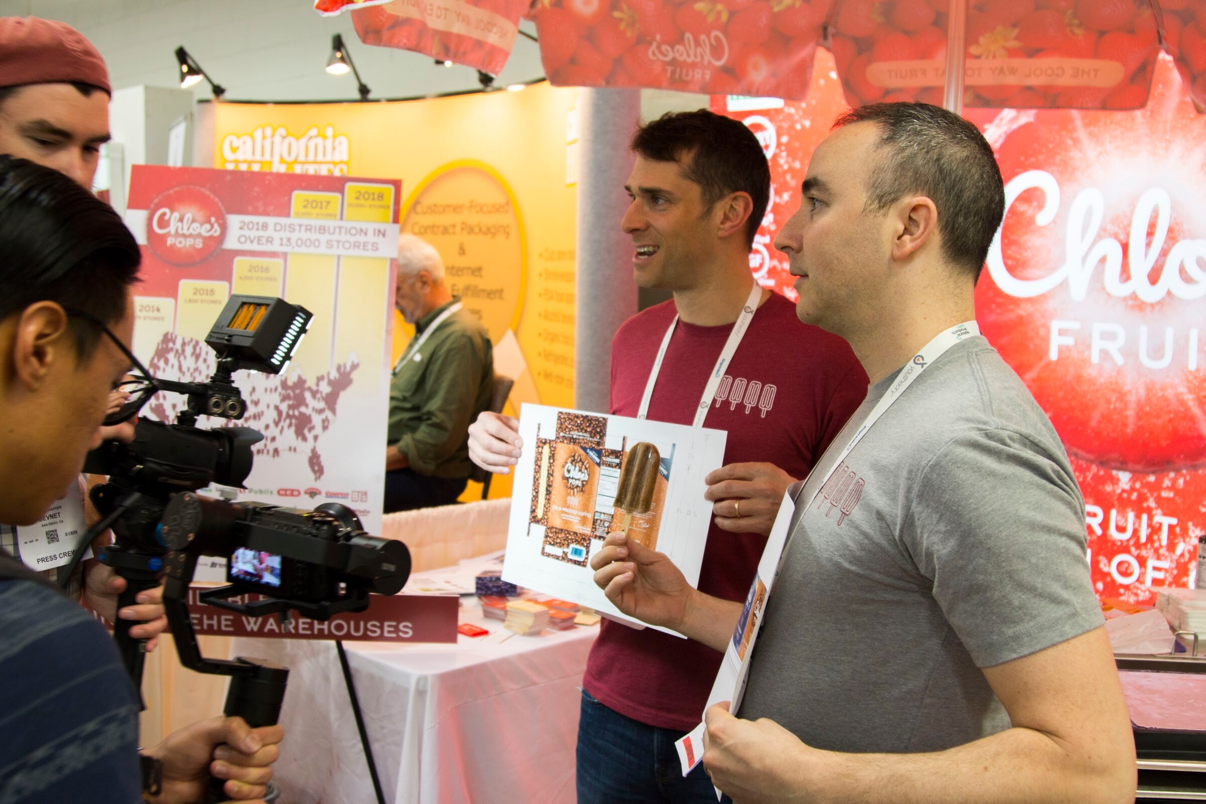 Two men in t-shirts holding a paper mockup of a popsicle package in front of a Chloe's Pops display, while a camera operator films at the left.