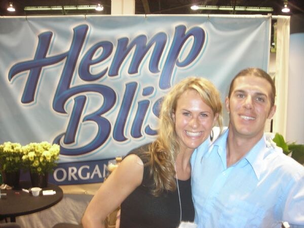 A smiling blonde woman and a man in a light blue shirt pose close together in front of a blue Hemp Bliss banner, with yellow flowers and a small table at left.