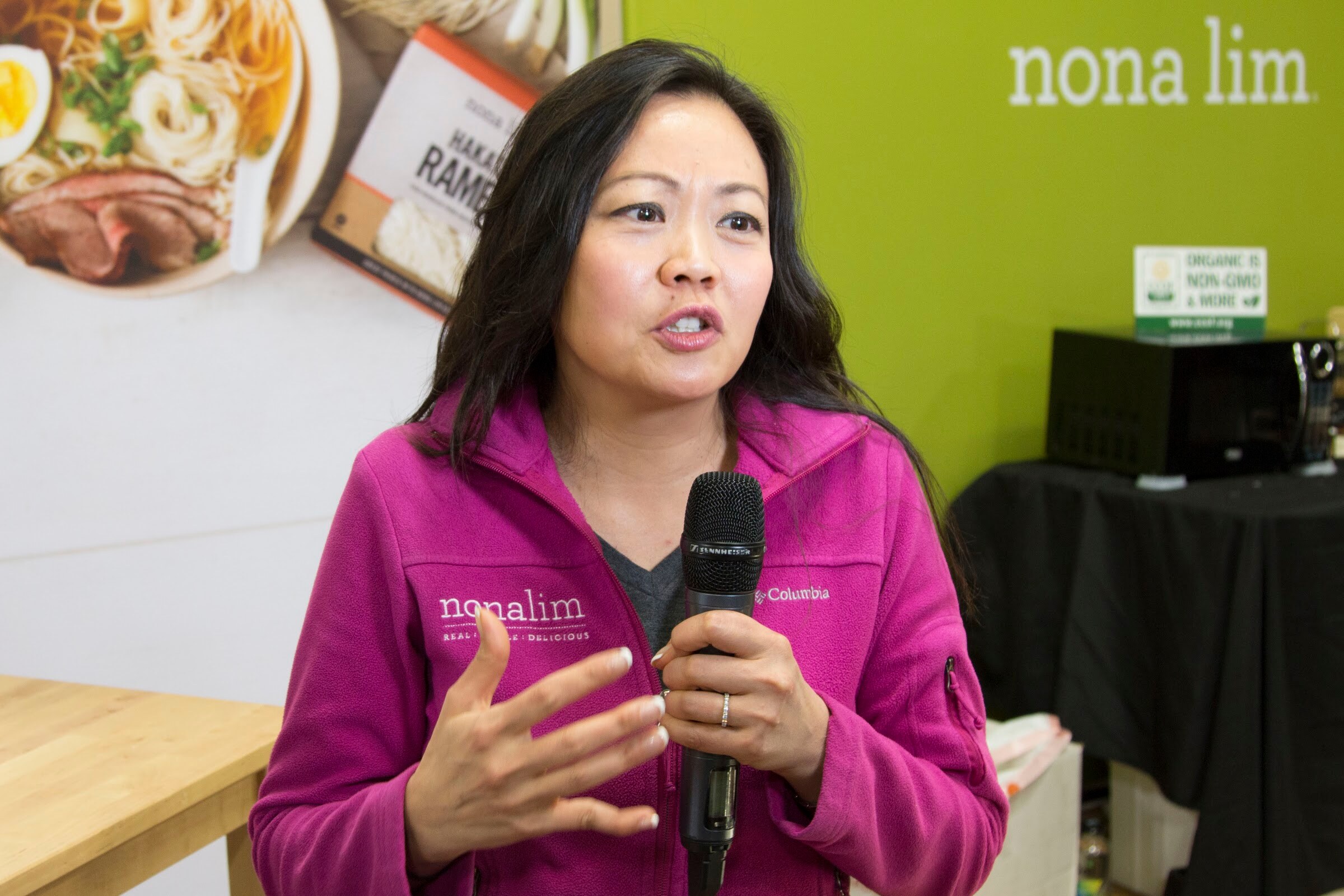 Woman in a magenta fleece jacket with the nona lim logo speaking into a microphone and gesturing, in front of a green wall.