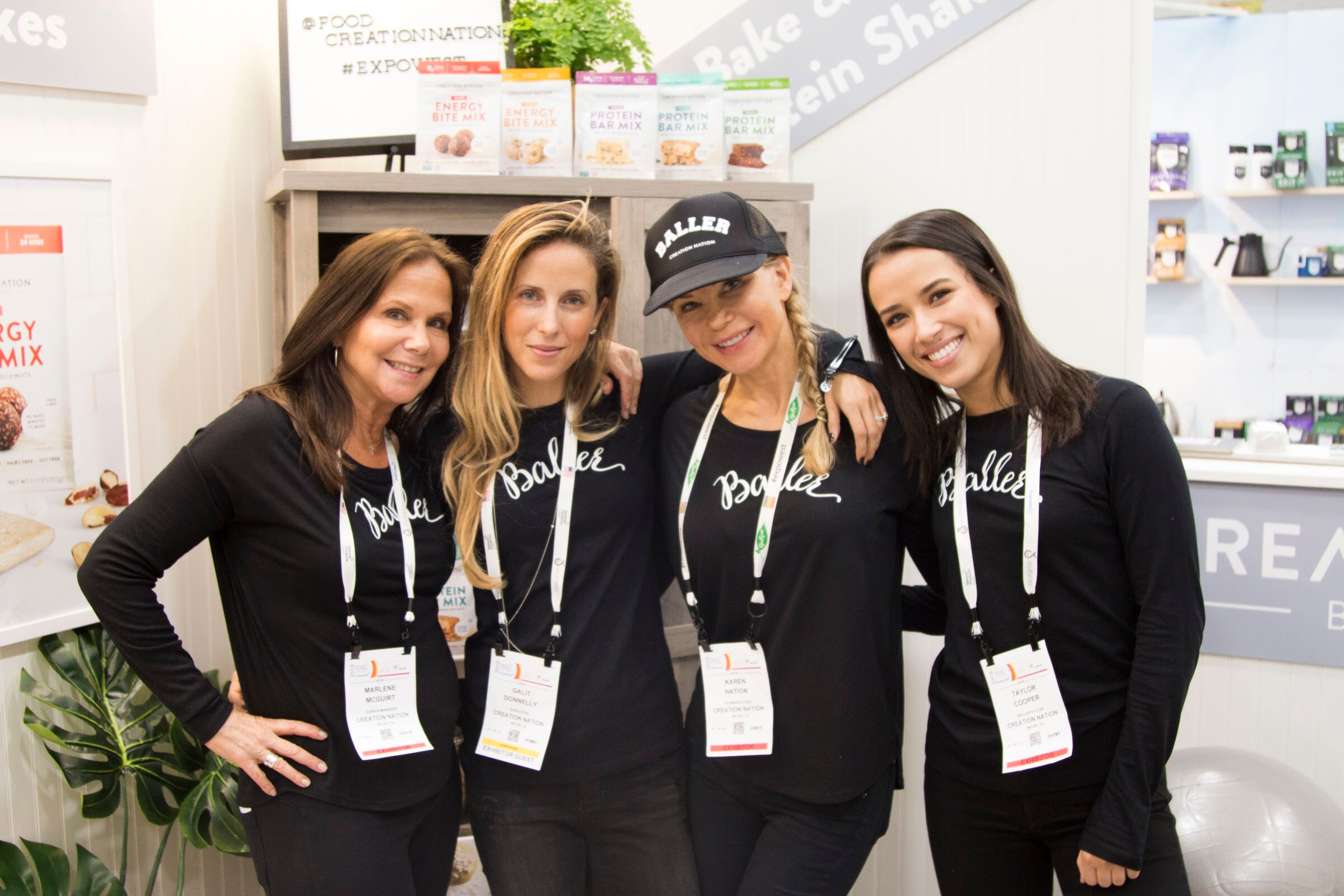 Four smiling women in black long-sleeve shirts printed with the word Baller, with lanyards, in front of shelves of protein products.