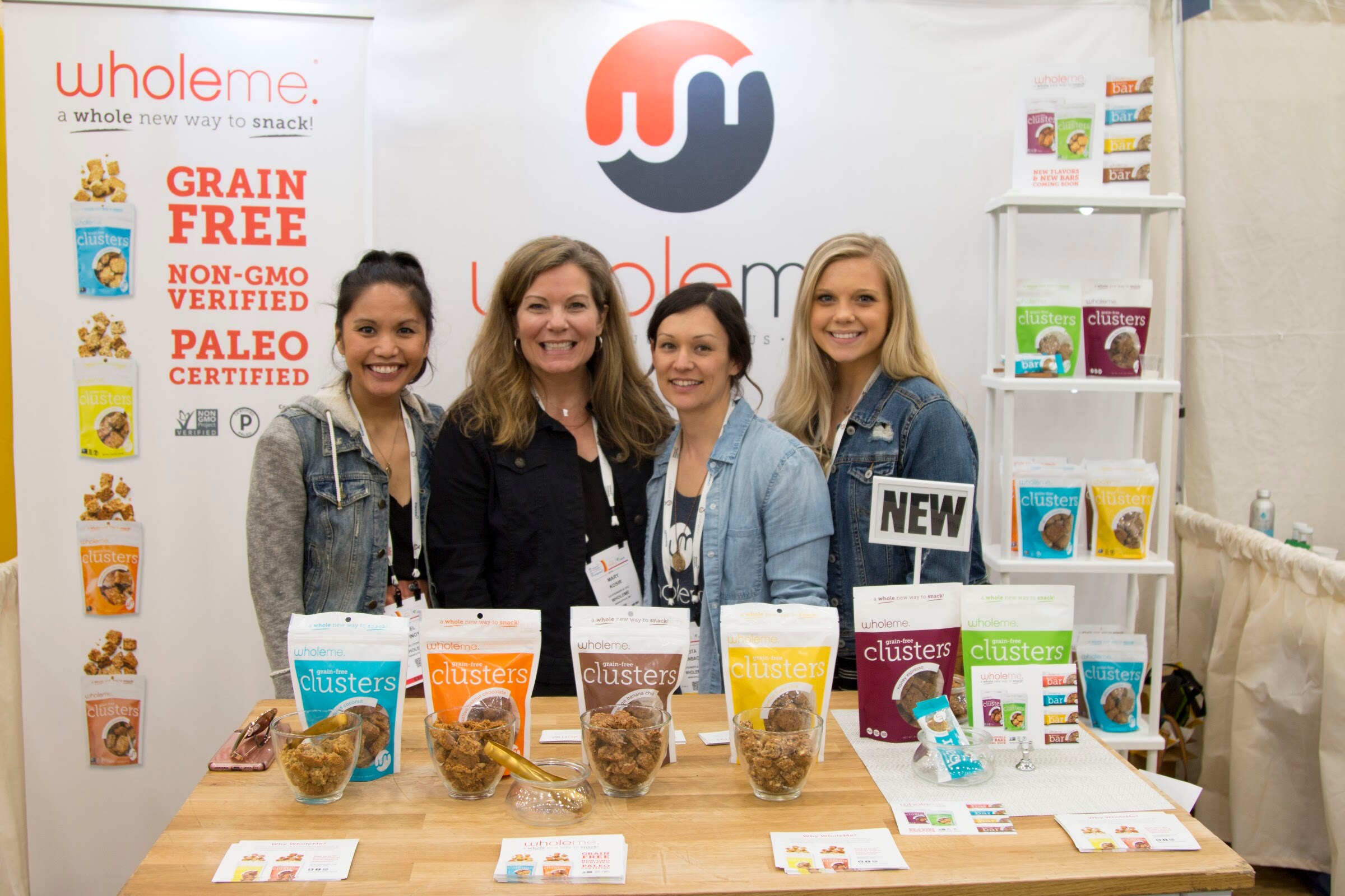 Four smiling women with lanyards behind a wooden counter with jars and bags of wholeme clusters and a Grain Free, Paleo banner.