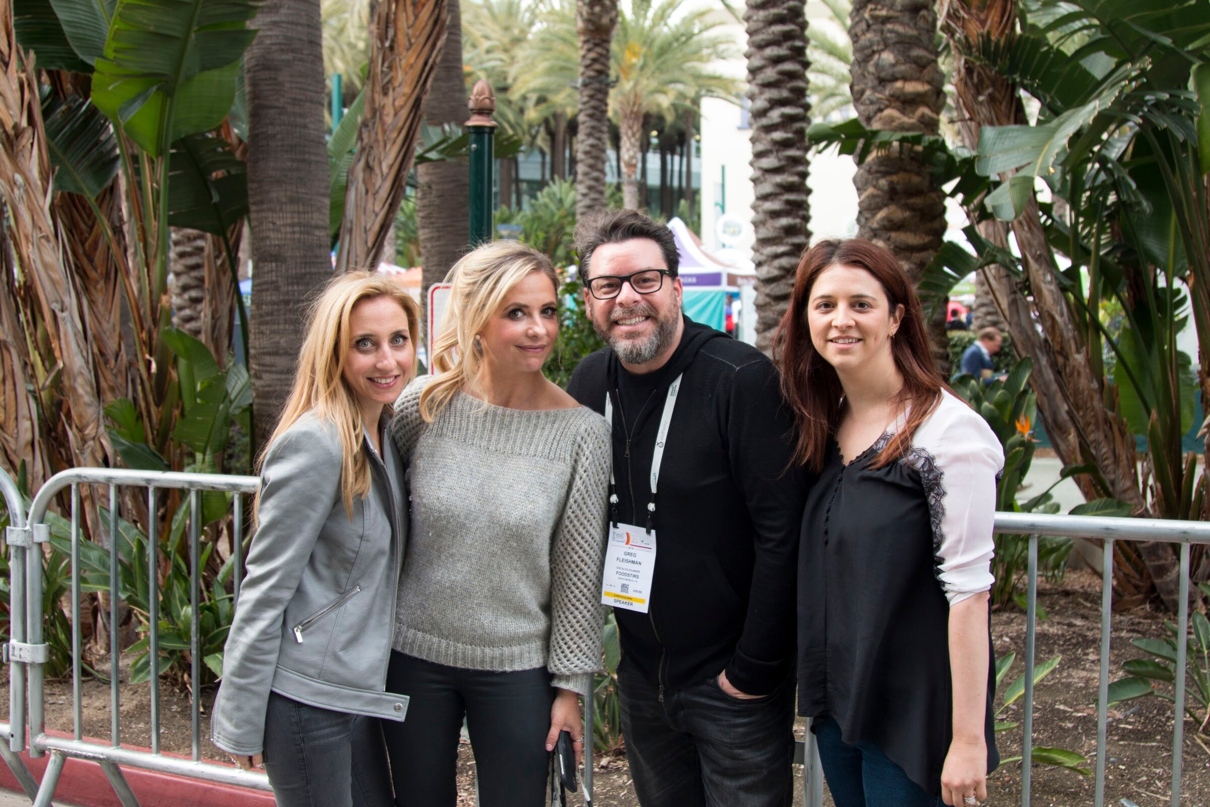 Four people standing close together with lanyards in front of palm trees and a metal crowd barrier.