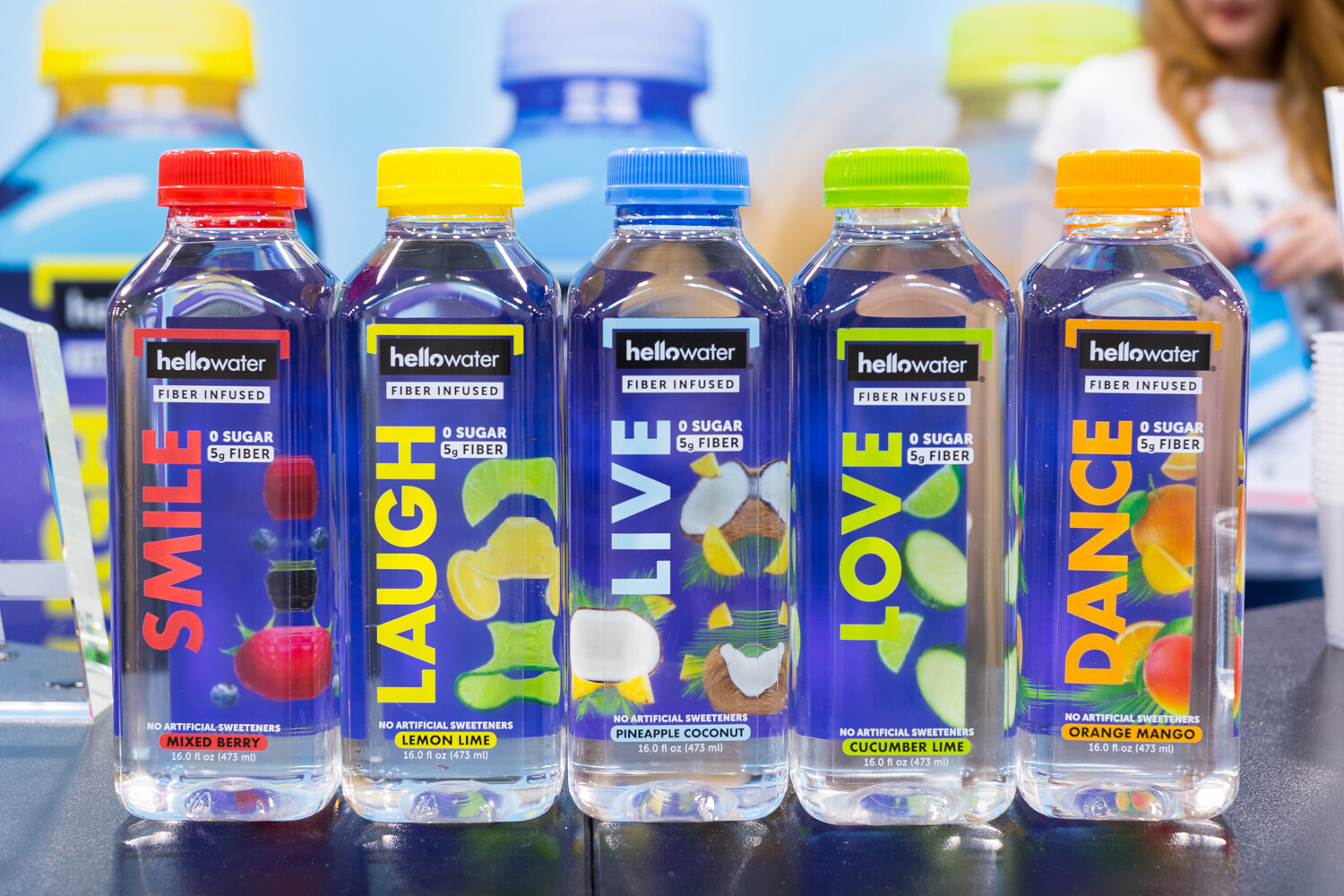 Five hellowater bottles with colored caps and the words smile, laugh, live, love, dance, with fruit graphics.